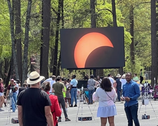 A photo of a crowd of people standing in front of a giant projection screen depicting the Moon's shadow passing in front of the Sun